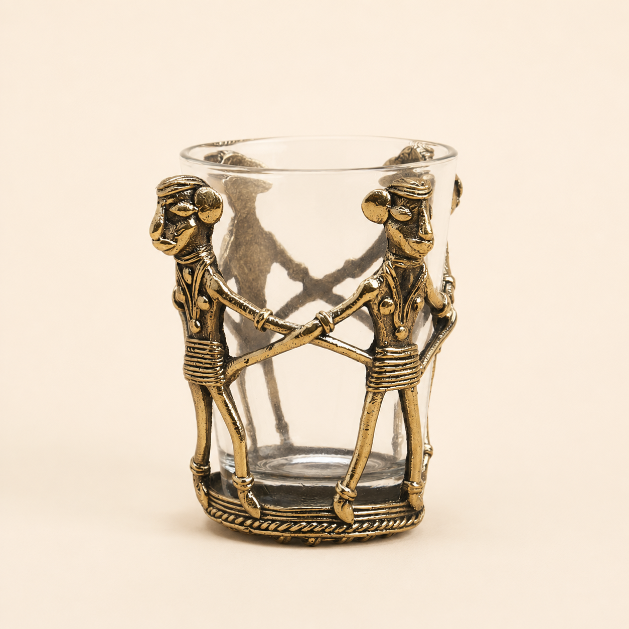 Dhokra brass tea holder with linked tribal figures and clear glass on an ivory studio background