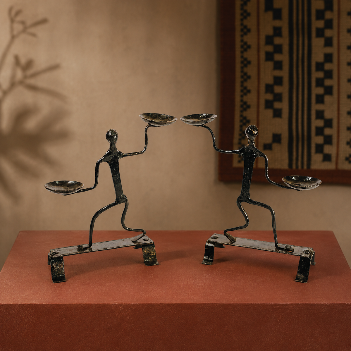 Two handcrafted Bastar iron candle holder figures displayed on an ochre gallery plinth